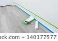 Home renovation scene featuring paint roller, tray, and painter's tape on freshly prepared floor with white walls and door frame visible 140177577