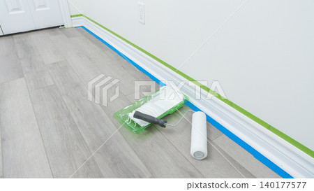 Home renovation scene featuring paint roller, tray, and painter's tape on freshly prepared floor with white walls and door frame visible Home renovation scene featuring paint roller, tray, and painter's tape on freshly prepared floor with white walls and door frame visible 140177577