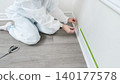 Person in protective suit applies painter's tape along baseboard in a home renovation project, scissors placed nearby on the floor 140177578