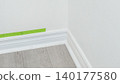 Corner of a room with freshly painted white walls, wooden floor, and green painter's tape applied along the baseboard for clean edges 140177580