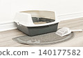 Cat litter box with a scoop placed on a gray mat, positioned on a wooden floor against a white wall in a clean indoor setting 140177582