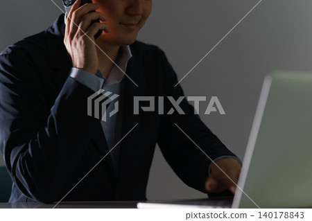 網路犯罪的畫面:一名身穿西裝的男子,在背光的映襯下,顯得身分更加隱蔽,正在操作電腦和智慧型手機。 網路犯罪的畫面:一名身穿西裝的男子,在背光的映襯下,顯得身分更加隱蔽,正在操作電腦和智慧型手機。 140178843
