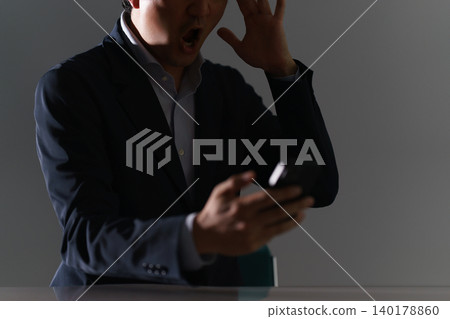 畫面中,一名三十多歲、身穿西裝、面容模糊的男子,在光線昏暗、背光的房間裡看著他的智慧型手機,嘴巴大張,一臉震驚。 畫面中,一名三十多歲、身穿西裝、面容模糊的男子,在光線昏暗、背光的房間裡看著他的智慧型手機,嘴巴大張,一臉震驚。 140178860
