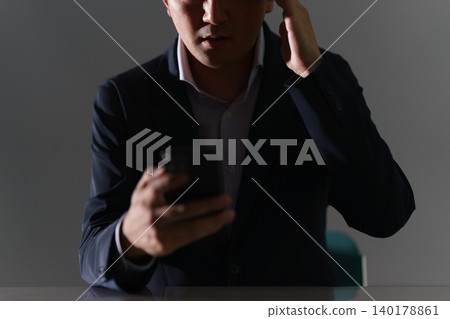 A man in a suit, whose face is obscured, sits in a dimly lit room with his smartphone in his hand, clutching his head in panic, having been scammed. A man in a suit, whose face is obscured, sits in a dimly lit room with his smartphone in his hand, clutching his head in panic, having been scammed. 140178861