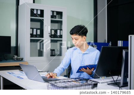 Asian businessman reviewing reports and working on laptop in a bright daytime office, highlighting productivity, business analysis, corporate workflow, and success. 140179685