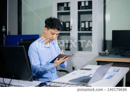 Asian businessman reviewing reports and working on laptop in a bright daytime office, highlighting productivity, business analysis, corporate workflow, and success. 140179686