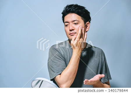 A summer image of a Japanese man in his 30s looking in a mirror, touching his cheek, and taking care of his skin, protecting himself from UV rays and sunburn. 140179844