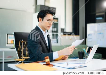 Professional Asian lawyer reading legal documents in modern office with gavel and scales on desk. Professional Asian lawyer reading legal documents in modern office with gavel and scales on desk. 140180046