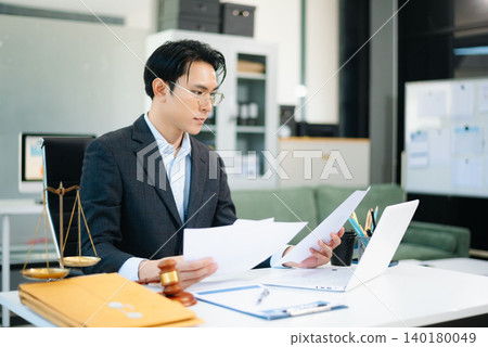 Professional Asian lawyer reading legal documents in modern office with gavel and scales on desk. Professional Asian lawyer reading legal documents in modern office with gavel and scales on desk. 140180049