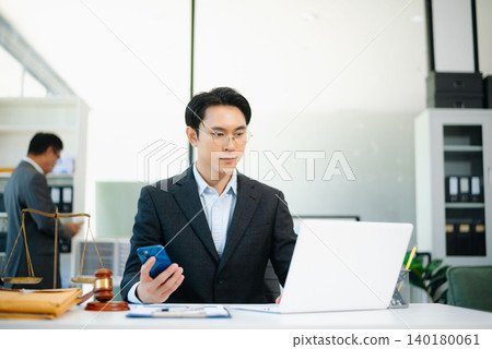 Asian lawyer in suit talking on phone while working on laptop with gavel and scales of justice on desk. Asian lawyer in suit talking on phone while working on laptop with gavel and scales of justice on desk. 140180061