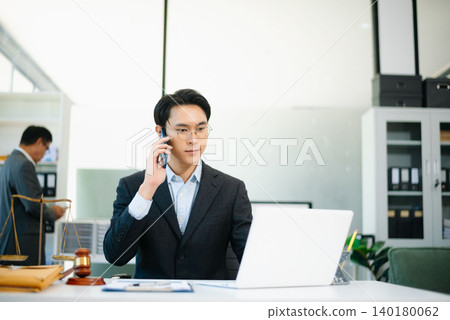 Asian lawyer in suit talking on phone while working on laptop with gavel and scales of justice on desk. Asian lawyer in suit talking on phone while working on laptop with gavel and scales of justice on desk. 140180062