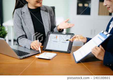 Two professional women in suits reviewing financial charts and analytics on paper and tablet in office. Business analysis, planning, and corporate strategy concept. Two professional women in suits reviewing financial charts and analytics on paper and tablet in office. Business analysis, planning, and corporate strategy concept. 140180601