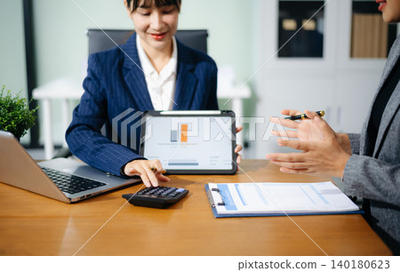 Two professional women in suits reviewing financial charts and analytics on paper and tablet in office. Business analysis, planning, and corporate strategy concept. Two professional women in suits reviewing financial charts and analytics on paper and tablet in office. Business analysis, planning, and corporate strategy concept. 140180623