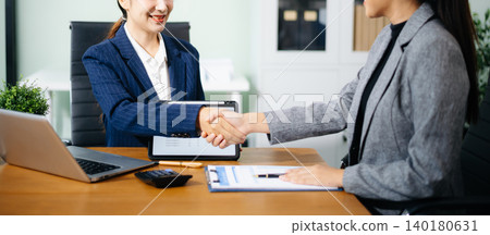 Two female professionals shake hands over financial reports and contract papers in an office, symbolizing successful business agreement, teamwork, and corporate partnership. 140180631