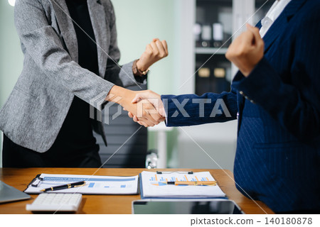 Two female professionals shake hands over financial reports and contract papers in an office, symbolizing successful business agreement, teamwork, Two female professionals shake hands over financial reports and contract papers in an office, symbolizing successful business agreement, teamwork, 140180878