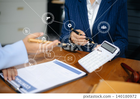 Two female professionals in business suits discussing a legal contract at office desk with gavel, folders, and calculator. Law, finance, and legal consultation theme. Two female professionals in business suits discussing a legal contract at office desk with gavel, folders, and calculator. Law, finance, and legal consultation theme. 140180972