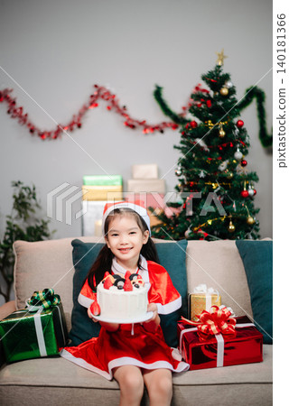 Christmas scene with a smiling young girl in Santa attire holding gifts and a cake, surrounded by holiday decor 140181366