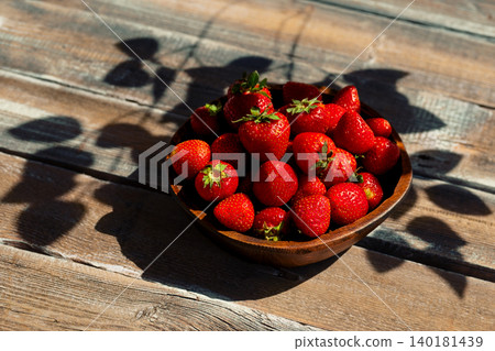 Fresh strawberries in wooden bowl on rustic table with shadows in afternoon light 140181439