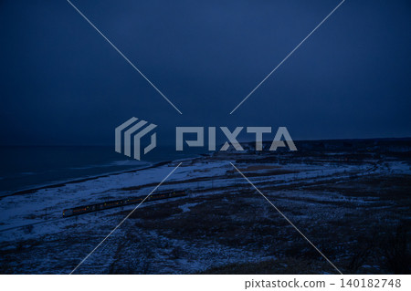 A coastal view from the hills of Shakubetsu, bathed in the blue light of the day. The snowfields, coastline, and train lights create a striking winter scene in Kushiro, Hokkaido. A coastal view from the hills of Shakubetsu, bathed in the blue light of the day. The snowfields, coastline, and train lights create a striking winter scene in Kushiro, Hokkaido. 140182748