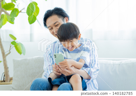 Family: Father and son sitting on the sofa looking at their smartphones. Family: Father and son sitting on the sofa looking at their smartphones. 140182785