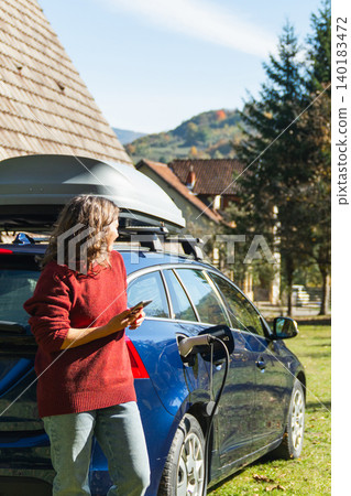Woman traveler with smartphone next to charging electric car on backyard of country house. 140183472