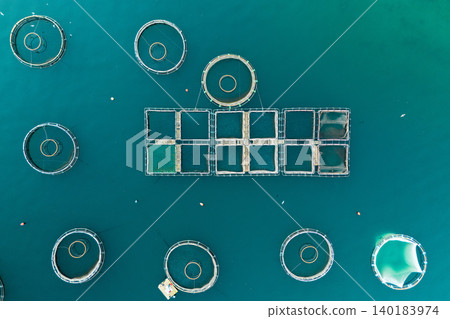 Aerial view of a fish farm located in a calm coastal bay, with floating cages arranged on the water surface. Aerial view of a fish farm located in a calm coastal bay, with floating cages arranged on the water surface. 140183974