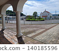 Weeds sprout from the grout lines of the tactile paving tiles in front of the station, and a white, Spanish-style bank building is visible between the arched columns. Weeds sprout from the grout lines of the tactile paving tiles in front of the station, and a white, Spanish-style bank building is visible between the arched columns. 140185596