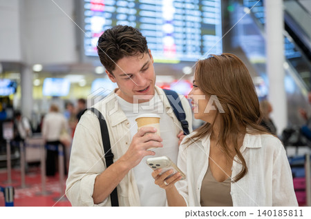 Happy asian woman girlfriend showing phone to white man boyfriend going on a trip waiting in airport Happy asian woman girlfriend showing phone to white man boyfriend going on a trip waiting in airport 140185811