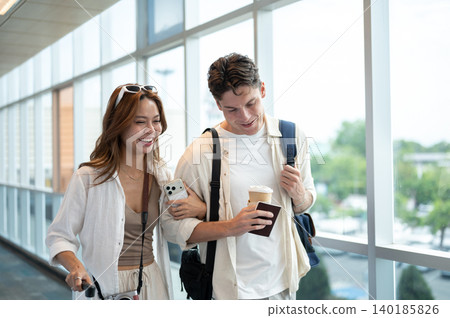 Smiling asian woman girlfriend linking arms talking to white man boyfriend while walking in airport. 140185826