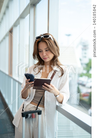Pretty asian woman looking at phone holding passport as standing at glass wall in airport's building Pretty asian woman looking at phone holding passport as standing at glass wall in airport's building 140185842