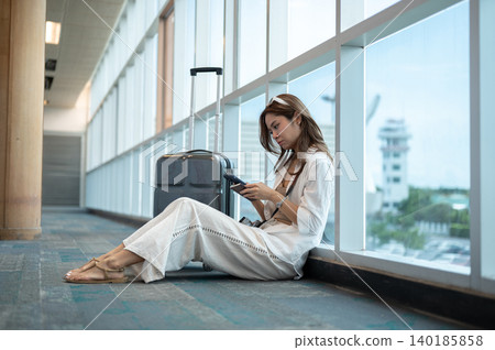 Bored asian woman holding passport looking at phone sitting on floor waiting in airport's building. Bored asian woman holding passport looking at phone sitting on floor waiting in airport's building. 140185858