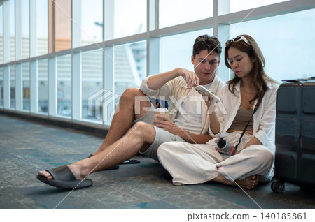 Asian woman girlfriend holding phone sitting with white man boyfriend an luggage on floor in airport Asian woman girlfriend holding phone sitting with white man boyfriend an luggage on floor in airport 140185861