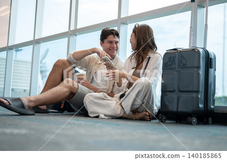 Asian woman girlfriend talking to white man boyfriend while sitting with luggage on floor in airport Asian woman girlfriend talking to white man boyfriend while sitting with luggage on floor in airport 140185865