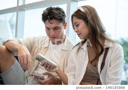 Pretty asian woman girlfriend looking at white man boyfriend's phone as sitting on floor in airport. 140185866