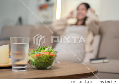 Close up of a glass of water and salad bowl on wooden table with blurred pregnant woman in the back. Close up of a glass of water and salad bowl on wooden table with blurred pregnant woman in the back. 140186116