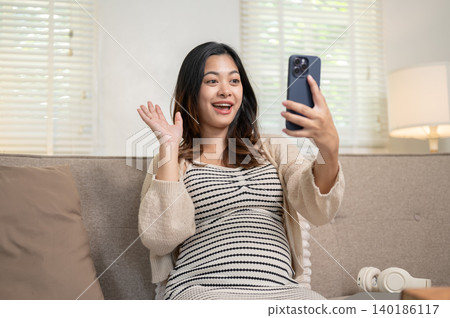 Surprised pregnant asian woman looking at phone on video call sitting on living room's sofa at home. Surprised pregnant asian woman looking at phone on video call sitting on living room's sofa at home. 140186117