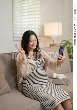 Happy pregnant asian woman looking at phone on video call while sitting on sofa at home living room. 140186118