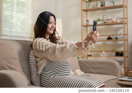 Happy pregnant asian woman holding phone taking picture while sitting on sofa in living room at home Happy pregnant asian woman holding phone taking picture while sitting on sofa in living room at home 140186119