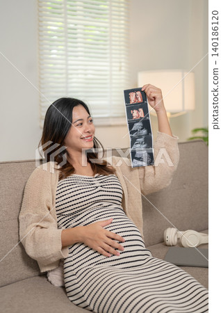 Pregnant asian woman holding ultrasound picture touching belly sitting on sofa at home's living room Pregnant asian woman holding ultrasound picture touching belly sitting on sofa at home's living room 140186120