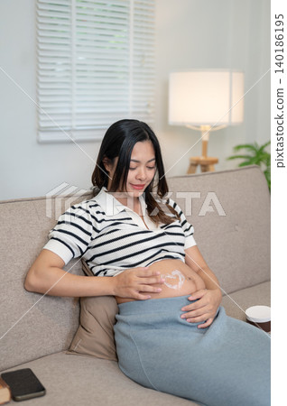 Pregnant asian woman applying lotion drawing heart on belly sitting on sofa in living room at home. Pregnant asian woman applying lotion drawing heart on belly sitting on sofa in living room at home. 140186195