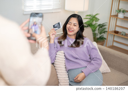 A friend taking photo of pregnant asian woman and ultrasound picture on phone in living room at home A friend taking photo of pregnant asian woman and ultrasound picture on phone in living room at home 140186207