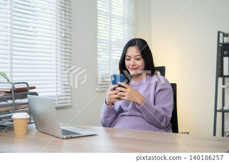 Pretty asian woman looking at phone beside laptop and coffee while sitting at wooden table in home. Pretty asian woman looking at phone beside laptop and coffee while sitting at wooden table in home. 140186257