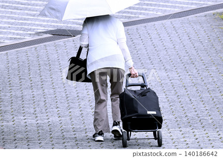 An elderly woman, holding a parasol and pulling a cart full of luggage, over a step in the cityscape of Yokohama, Japan. An elderly woman, holding a parasol and pulling a cart full of luggage, over a step in the cityscape of Yokohama, Japan. 140186642