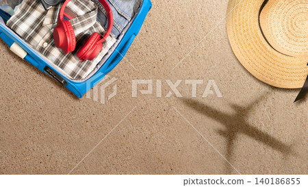 Top view of a packed blue suitcase, red headphones, and a straw hat on beach sand with a shadow of an airplane. Concept of summer vacation and travel planning Top view of a packed blue suitcase, red headphones, and a straw hat on beach sand with a shadow of an airplane. Concept of summer vacation and travel planning 140186855