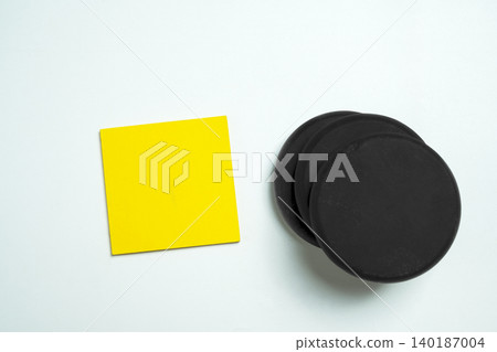 A bright yellow square sticky note placed next to a stack of black round magnets or clay discs on a clean white surface, offering a minimalist workspace mockup A bright yellow square sticky note placed next to a stack of black round magnets or clay discs on a clean white surface, offering a minimalist workspace mockup 140187004