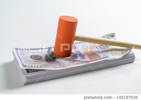 A wooden toy gavel mallet with an orange head rests on top of a stack of hundred US dollar bills, symbolizing legal costs, court fees, judgment, and auctions A wooden toy gavel mallet with an orange head rests on top of a stack of hundred US dollar bills, symbolizing legal costs, court fees, judgment, and auctions 140187036