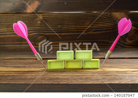 Two pink darts stuck in wood next to a stack of green rectangular blocks. Concept of strategy, business goals, and hitting targets on a rustic wooden background Two pink darts stuck in wood next to a stack of green rectangular blocks. Concept of strategy, business goals, and hitting targets on a rustic wooden background 140187047