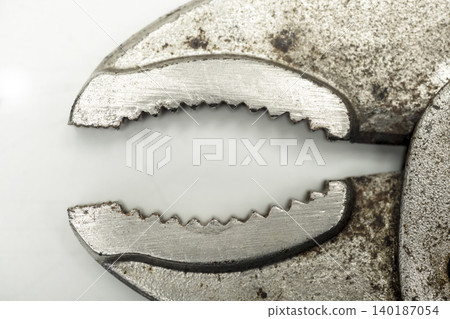 A macro shot focusing on the textured, worn, and slightly rusted metallic jaws and teeth of an old adjustable pair of pliers on a light background A macro shot focusing on the textured, worn, and slightly rusted metallic jaws and teeth of an old adjustable pair of pliers on a light background 140187054