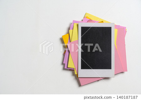 Top-down view of a blank instant photo frame on a stack of colorful pink and yellow paper notes over a white textured background. Ideal for mockups, memory concepts, and creative design 140187187