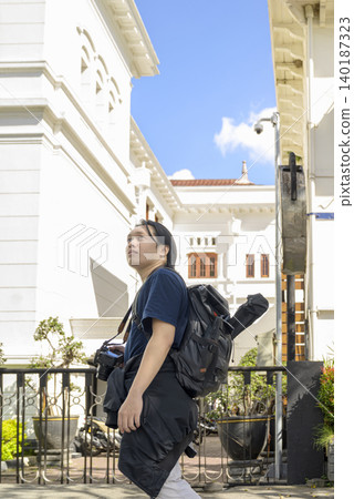 A young Indonesian southeast asian tourist carrying a backpack and camera walks along a street with colonial-style white buildings on a sunny day. Travel or vacation concept A young Indonesian southeast asian tourist carrying a backpack and camera walks along a street with colonial-style white buildings on a sunny day. Travel or vacation concept 140187323